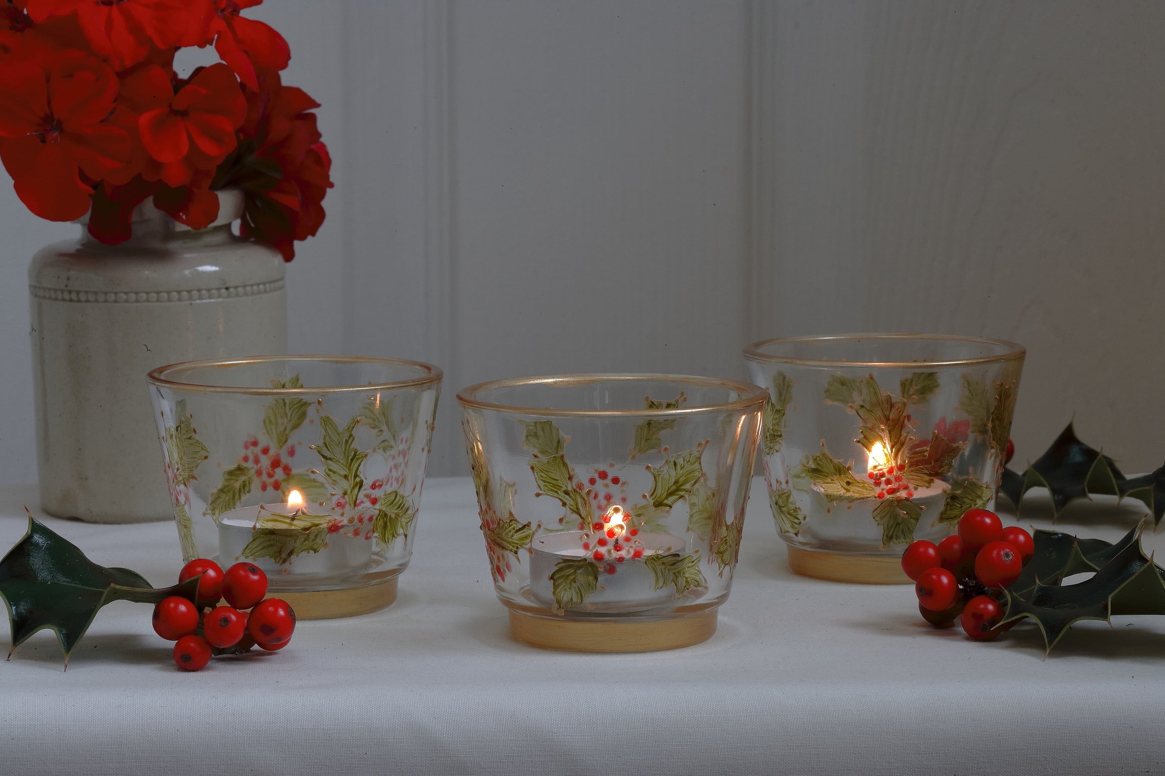 Decorative glass candle holders with floral designs on a white surface, surrounded by red berries and green leaves.