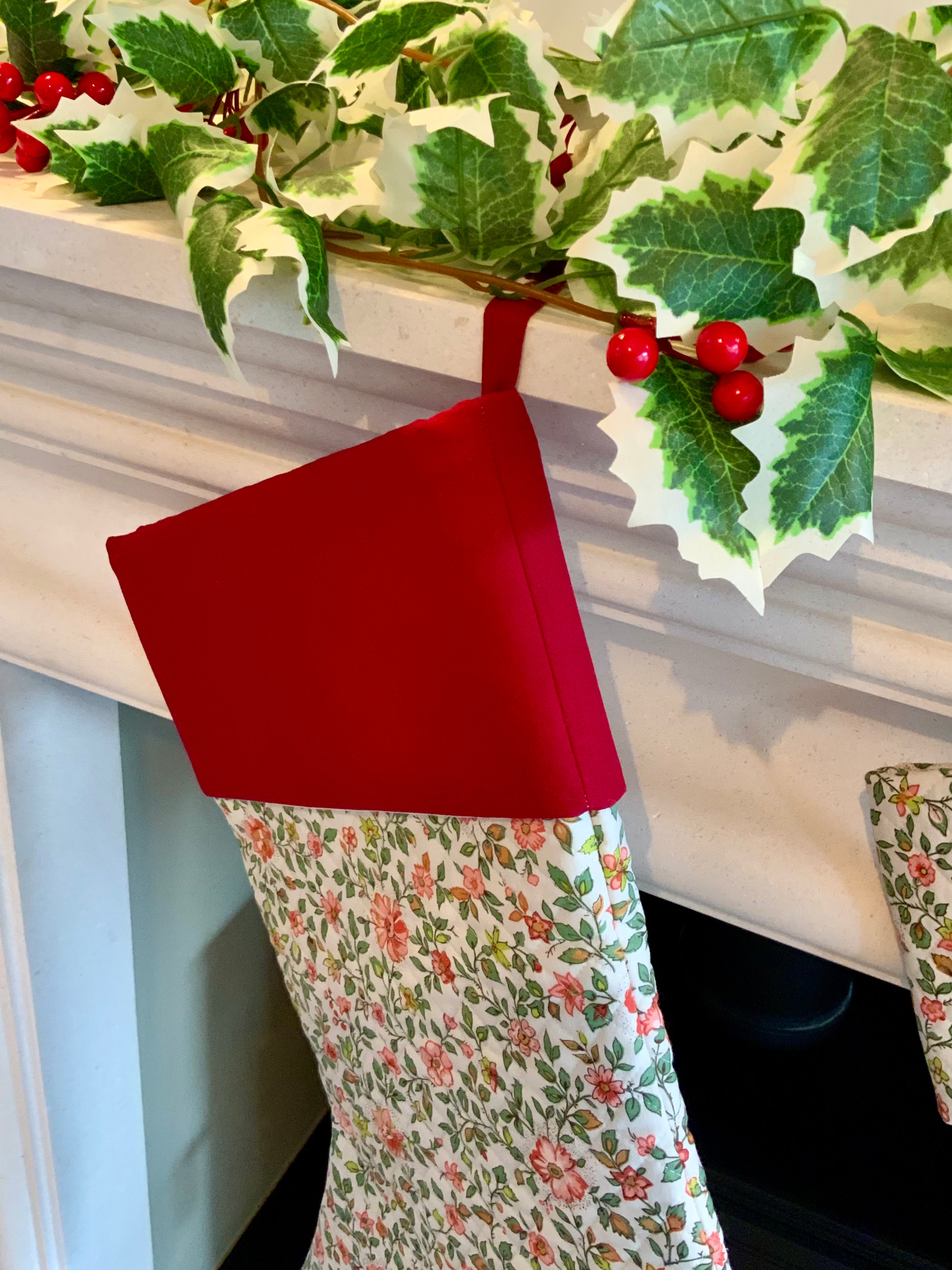 Red and floral-patterned Christmas stockings hanging on a fireplace with holly leaves and berries.