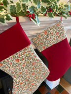 Two floral-patterned Christmas stockings on a fireplace with holly leaves and berries.