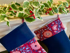 Decorative stockings with floral patterns hanging on a mantelpiece with green leafy branches.