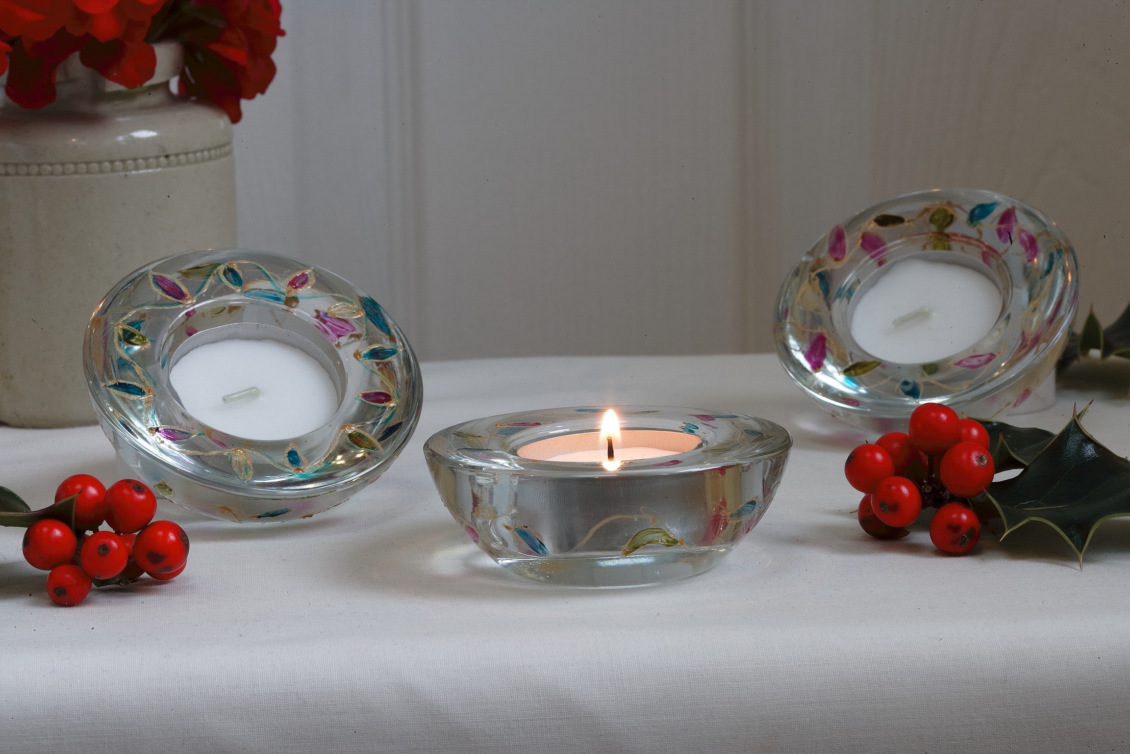 Decorative candle holders with candles on a white surface, surrounded by red berries and holly.