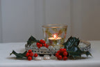 Decorative setup with a lit candle in a glass holder, surrounded by holly leaves and berries on a white surface.
