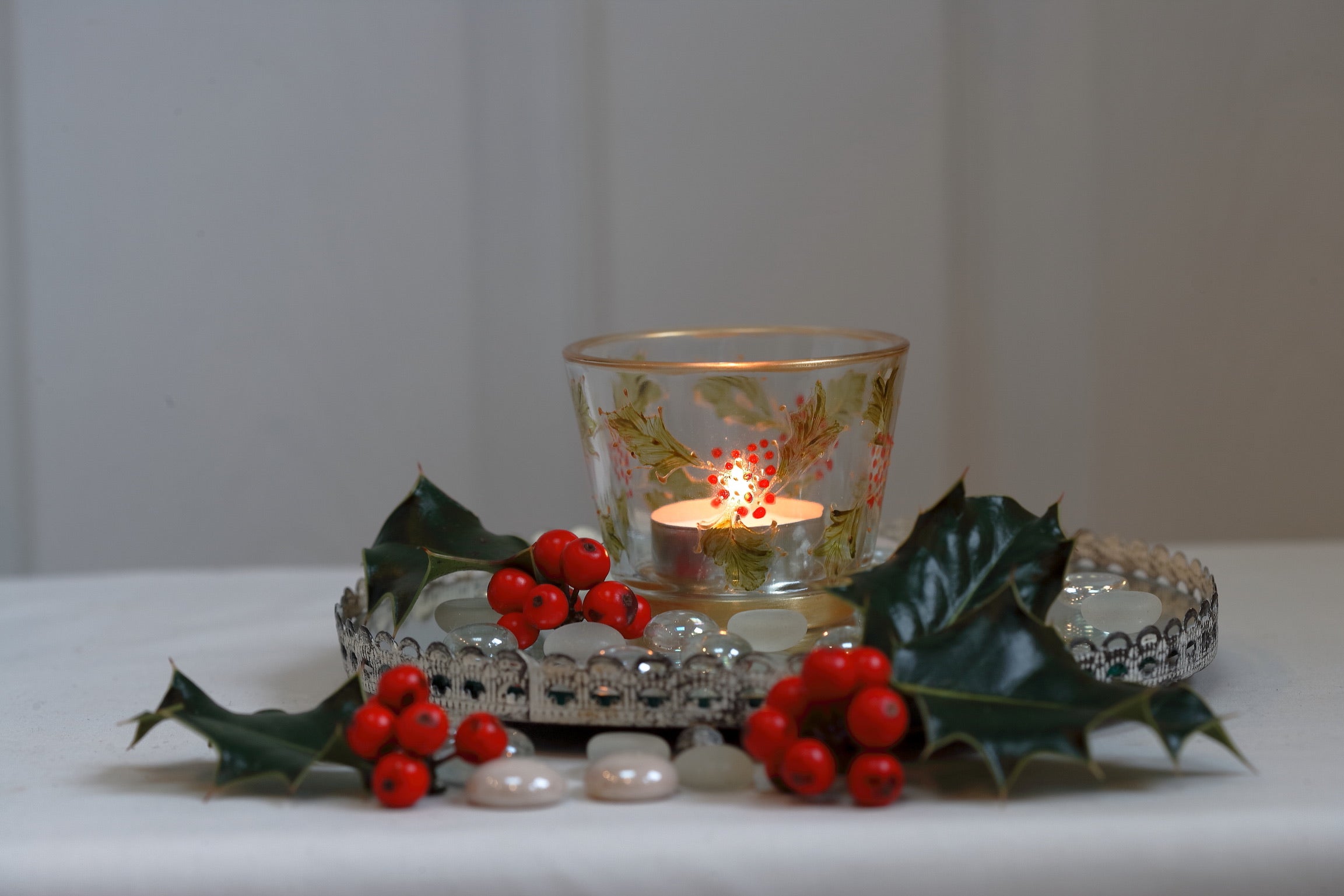 Decorative setup with a lit candle in a glass holder, surrounded by holly leaves and berries on a white surface.
