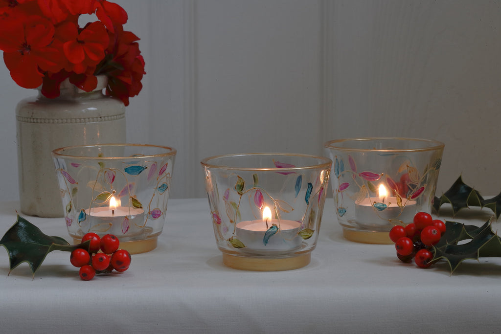 Three small glass candle holders with lit candles on a white surface, surrounded by red berries and green leaves.